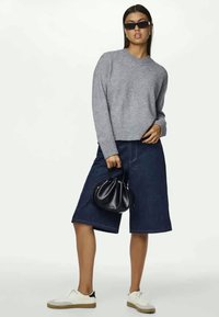 Grau gestrickter Pullover, mittellange blaue Jeansshorts, schwarz-weiße Sneaker, schwarze Handtasche und dunkle Sonnenbrille. Einfaches, lässiges Outfit.