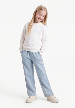Fille souriante aux longs cheveux blonds portant un sweat-shirt blanc, un pantalon large bleu clair avec un logo et des baskets beiges, debout les mains dans les poches.