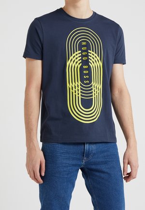 Man draagt een marineblauw T-shirt met een geel ovale afbeelding en de tekst "Hugo Boss", gecombineerd met een blauwe spijkerbroek tegen een effen achtergrond.