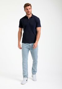 Mornarska polo majica z rebrastim ovratnikom in trakom s tremi gumbi, v kombinaciji s svetloblueskimi kavbojkami in belimi supergami, stoji ob enotnem ozadju.