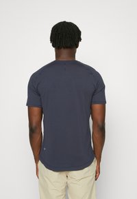 Marineblaues Kurzarmhemd mit Rundhalsausschnitt, weichem Material, abgerundetem Saum und subtilen Nahtdetails, getragen mit beigen Hosen, von hinten betrachtet.