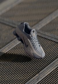 Šviesiai pilki sportiniai bateliai su tinklelio viršumi, tekstūruotais akcentais ir juodu "Nike" ženklu. Juose yra paminkštintas padas su išskirtiniu kulno dizainu.