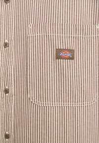 Brown and cream striped button-up shirt with a chest pocket and a Dickies logo patch. Made of fabric with a smooth texture.