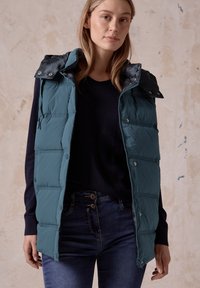 Teal vadderad väst med dragkedja fram och tryckknappar, med avtagbar huva och stickade paneler. Bärs över en marinblå tröja och jeans.