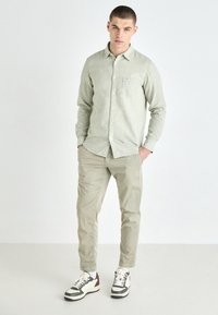 Light green button-up shirt with a chest pocket, paired with light beige pants and white sneakers featuring neutral accents. Smooth fabric texture.