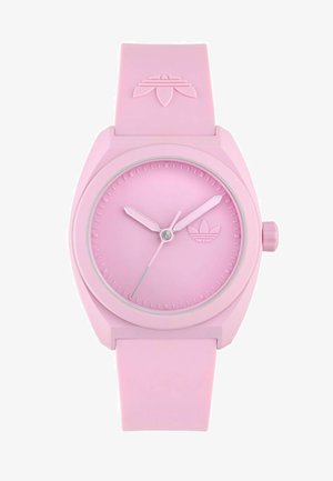 Rozā silikona pulkstenis ar apaļu seju, kontrastējošām gaiši rozā stundu un minūšu rādītājiem, un zīmola logo iegravēts skaitītājā.