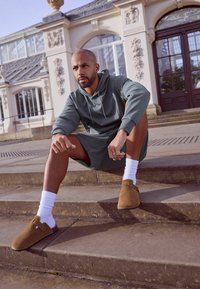 Brune ruskind slip-on sko med en spændeaccent, parret med hvide ankelsokker. Modellen har en grøn hoodie og matchende shorts på og sidder på stenskridt.