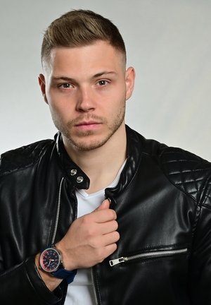 Black leather jacket with quilted shoulders, white t-shirt, blue watch with a round face and red accents; model has short, styled hair.