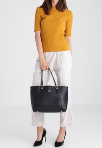 Černá kožená taška s dvojitými rukojeťmi, hladkým povrchem a předním detailem loga. Nese ji osoba v žlutém žebrovaném topu a bílých kalhotách.