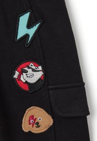 Schwarzer Stoff mit drei bestickten Aufnähern: einem blauen Blitz, einem runden roten Abzeichen mit einer Figur und einem Bärenkopf in Braun.