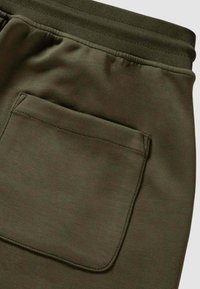 Tessuto verde scuro con una grande tasca rettangolare ed elastico in vita, probabilmente parte di pantaloni casual o da ginnastica.