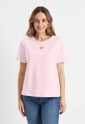 Vrouw met lichtbruin haar draagt een lichtroze t-shirt met een klein zilveren hartontwerp en blauwe spijkerbroek, glimlachend tegen een witte achtergrond.
