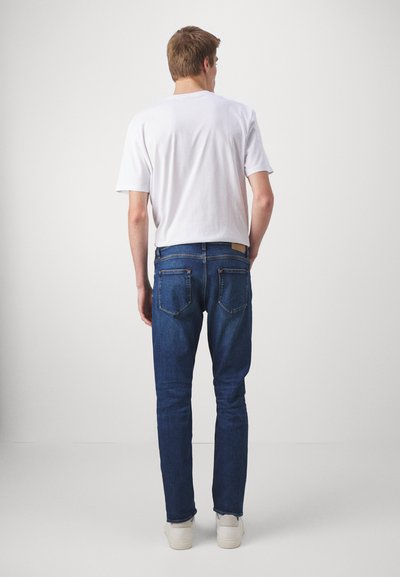 Blå denim jeans med rett benkutt, med to baklommer og et brunt lærmerke i midjen. Kombinert med en hvit t-skjorte.