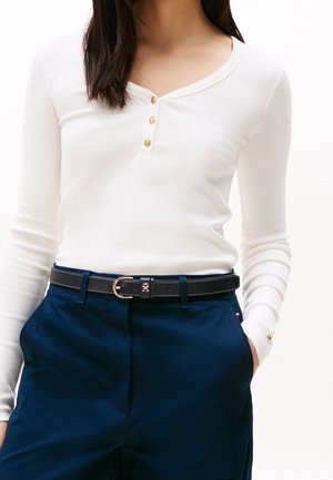 Femme portant un haut blanc à manches longues boutonné, rentré dans un pantalon bleu marine avec une ceinture noire, mains dans les poches, sur un fond uni.