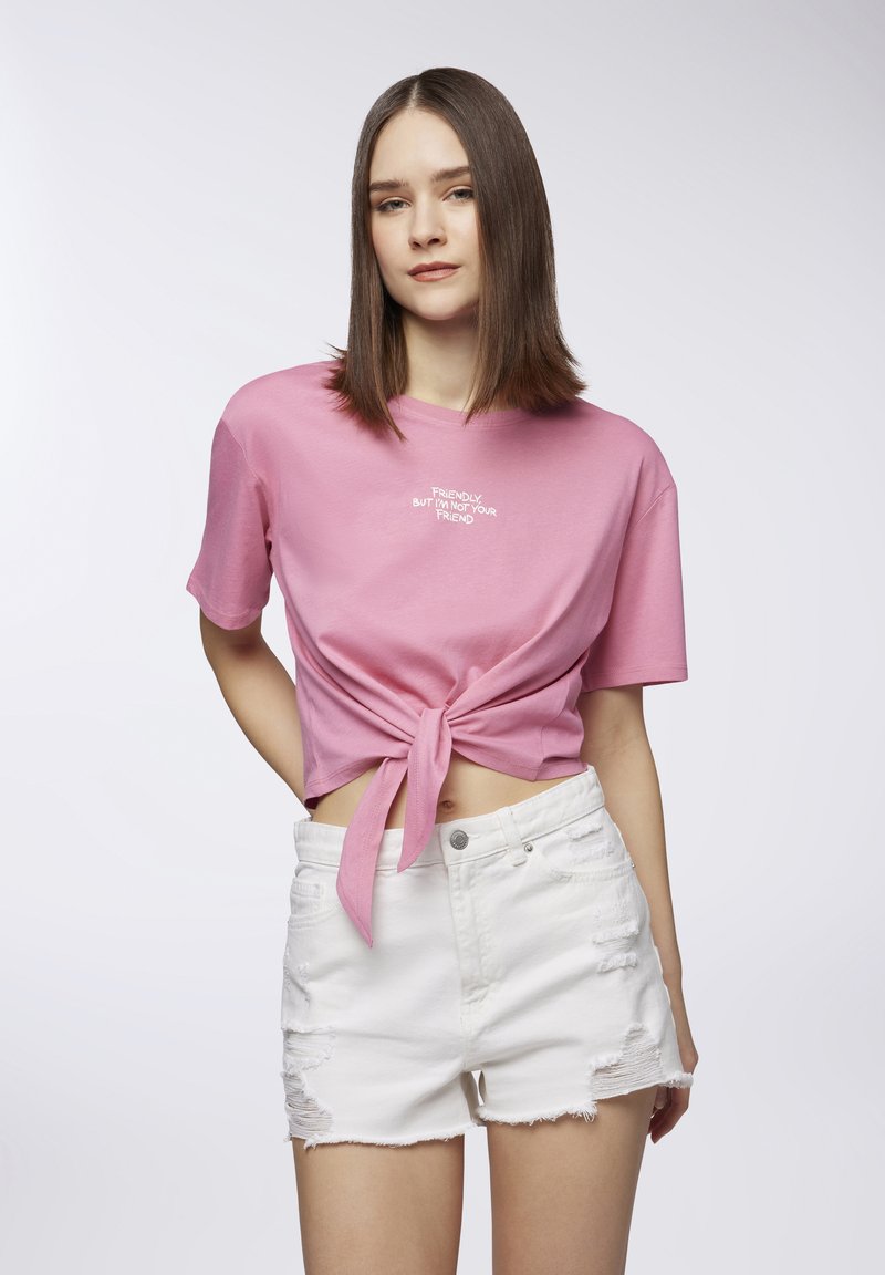 T-shirt in cotone rosa con dettaglio annodato in vita, caratterizzato da testo ricamato bianco. Abbinato a pantaloni corti in denim bianco in stile usurato.