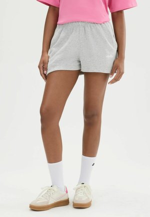 Persona que lleva puestos pantalones cortos grises con cintura elástica, calcetines blancos hasta el tobillo, zapatillas crema con suelas de goma y una camiseta rosa, de pie sobre un fondo blanco.