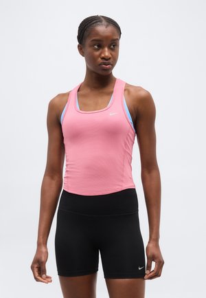 Atleetlik naine punutud soenguga kannab roosat varrukateta särk sinise spordirinnahoidja ja mustade vormis pükstega taustal tavalise tausta ees.