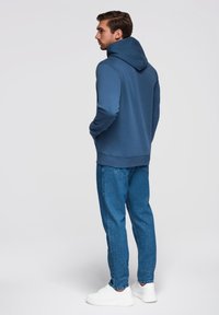 Felpa blu con cappuccio regolabile, abbinata a jeans in denim azzurro e sneakers bianche. Texture liscia e vestibilità rilassata.