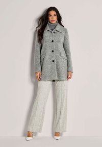 Vrouw in een grijze, knoopbare wollen jas over een grijze coltrui, met lichtgekleurde, breedpijped texturaal pantalons en witte hakken.