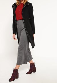 Manteau long noir avec fermeture éclair argentée, chemise rouge boutonnée, pantalons gris à jambes larges, et bottines bordeaux à motifs avec talons épais.