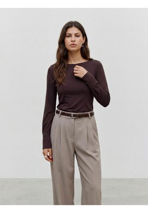 Femme aux longs cheveux foncés porte un haut moulant à manches longues marron foncé et un pantalon plissé beige taille haute avec une ceinture sombre, debout contre un mur uni.