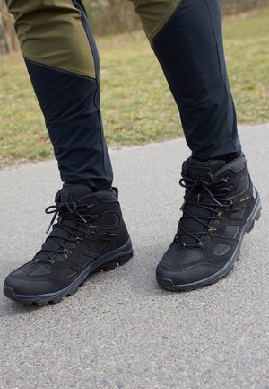 Personne portant des chaussures de randonnée noires et un pantalon de plein air noir et olive, debout sur un chemin pavé avec de l'herbe en arrière-plan.