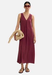 Bordowa, bezrękawnikowa sukienka maxi z dekoltem w kształcie litery V, z marszczeniami, noszona z plecioną beżową torbą i czarnymi sandałami na paski. Czerwone okulary przeciwsłoneczne dopełniają całość.