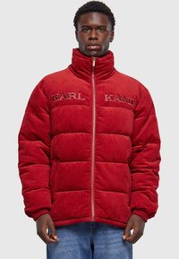 Rød corduroy pufferjakke med høj krave, der har quiltede sektioner og broderet "KARL KANI" branding på forsiden. Lynlåslukning.