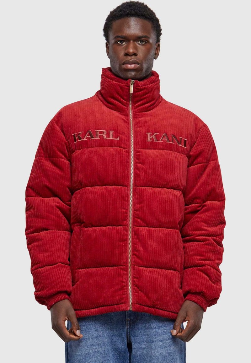Rød corduroy pufferjakke med høj krave, der har quiltede sektioner og broderet "KARL KANI" branding på forsiden. Lynlåslukning.