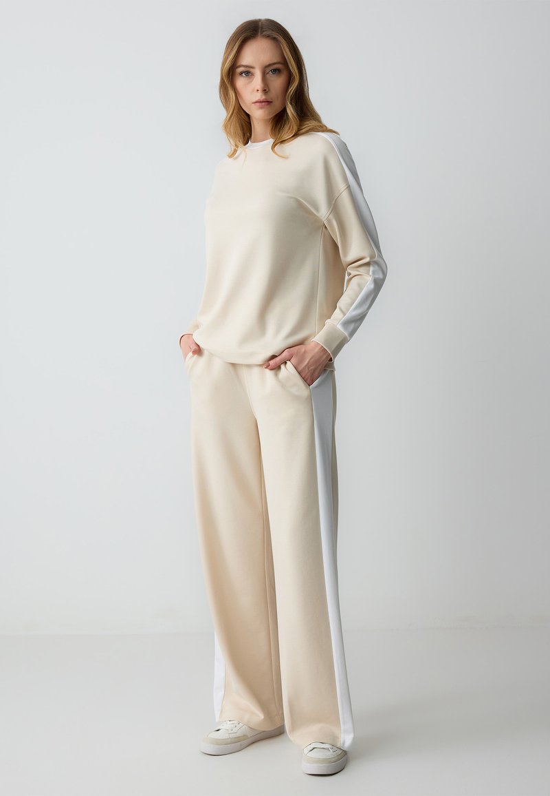 Beige sweatshirtoutfit gemaakt van zachte stof; lange mouwen en broek met witte zijkanten; relaxte pasvorm; zakken op de broek; witte sneakers.