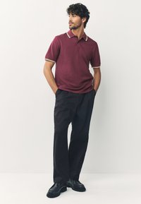 Bordeauxrotes Polohemd mit kontrastierendem, gestreiftem Kragen, kombiniert mit schwarzen Hosen und schwarzen Schuhen. Der Stoff wirkt glatt und der Schnitt maßgeschneidert.