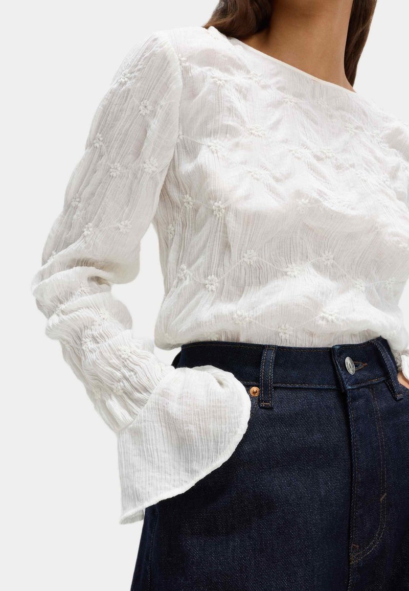 Blusa bianca con texture e motivi ricamati, maniche a sbuffo e scollatura rotonda, abbinata a jeans a vita alta in denim scuro.