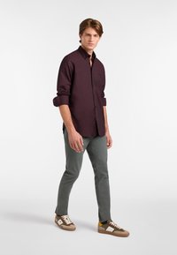 Giovane uomo che cammina, indossando una camicia a bottoni di un viola scuro, pantaloni grigi e sneakers beige con accenti bianchi e gialli.