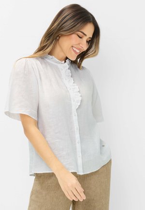 Frau lächelnd und nach unten schauend, trägt eine weiße, geknöpfte Bluse mit Rüschen und beige Hosen vor einem schlichten weißen Hintergrund.