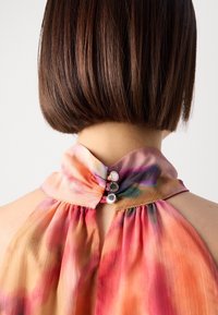 Femme aux cheveux bruns courts portant un haut sans manches multicolore fermé dans le dos par trois petits boutons argentés.