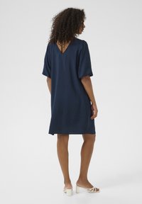 Navy blue short-sleeve dress with a V-back neckline, relaxed fit, textured fabric, and knee-length design. Worn with white block-heeled sandals.