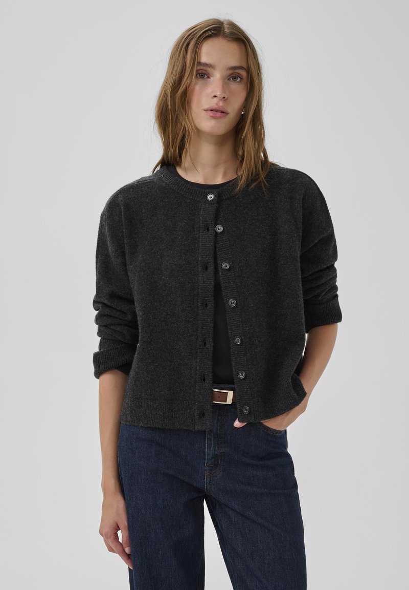 Cárdigan de lana gris oscuro con escote redondo, cierre de botones y ajuste ligeramente holgado; combinado con vaqueros de mezclilla oscura.