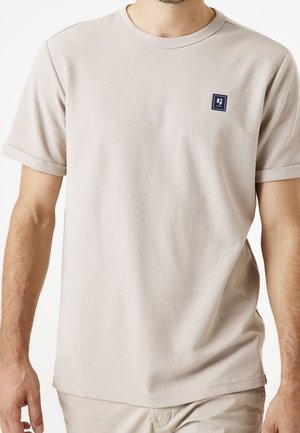 Mężczyzna ubrany w beżowy, krótki rękaw, teksturowany t-shirt z małym granatowym kwadratowym logo na lewej piersi.