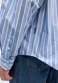 Chemise à manches longues rayée bleu et blanc avec poignets boutonnés, une petite étiquette bleue "Hugo", portée avec un jean en denim bleu.
