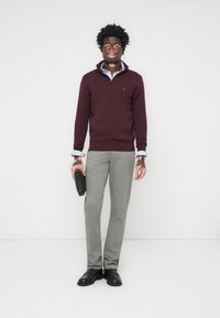 Bordeaux ritsweater over een blauw gestreept shirt, grijze broek, houdt een zwart voorwerp vast. Schoenen zijn zwart en het haar is gestyled in strakke krullen.
