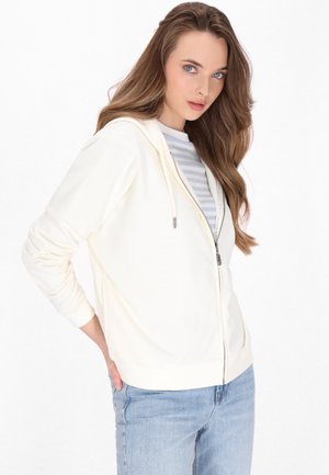White zip-up hoodie made from soft fabric, featuring a hood, front pockets, and ribbed cuffs, layered over a gray and white striped shirt.