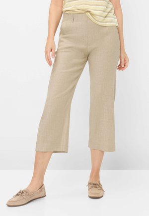 Frau trägt beige weit geschnittene, gekürzte Hosen, gestreiftes gelbes Oberteil und beige Schnürslipper und steht vor einem weißen Hintergrund.