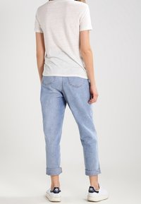 Žena v světle modrých zkrácených džínách, bílé košili s krátkým rukávem a bílých teniskách Adidas s námořnickými detaily, stojí zády k fotoaparátu.