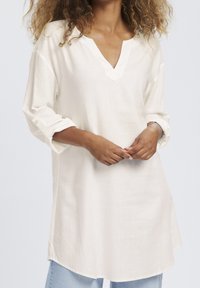 Biała lniana tunika z długim rękawem, dekoltem w serek, podwiniętymi mankietami oraz luźnym krojem. Miękka w dotyku i z lekko zaokrąglonym dołem dla wygody.