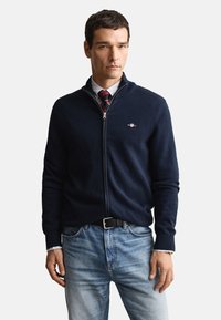 Marineblå genser med glidelås laget av strikket stoff, med en liten logo og ribbet krage, kombinert med lyseblå jeans.