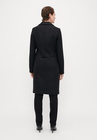 Μαύρο μακρύ παλτό με εφαρμοστή σχεδίαση, με ελαφρά θεακό λαιμό, και κλείσιμο με κουμπί, συνδυασμένο με μαύρα εφαρμοστά παντελόνια και ankle boots.