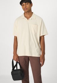 Hellbeige Polohemd mit einem lockeren Schnitt, ausgestattet mit einer Brusttasche. Kombiniert mit braunen Hosen und einer kleinen schwarzen Umhängetasche.