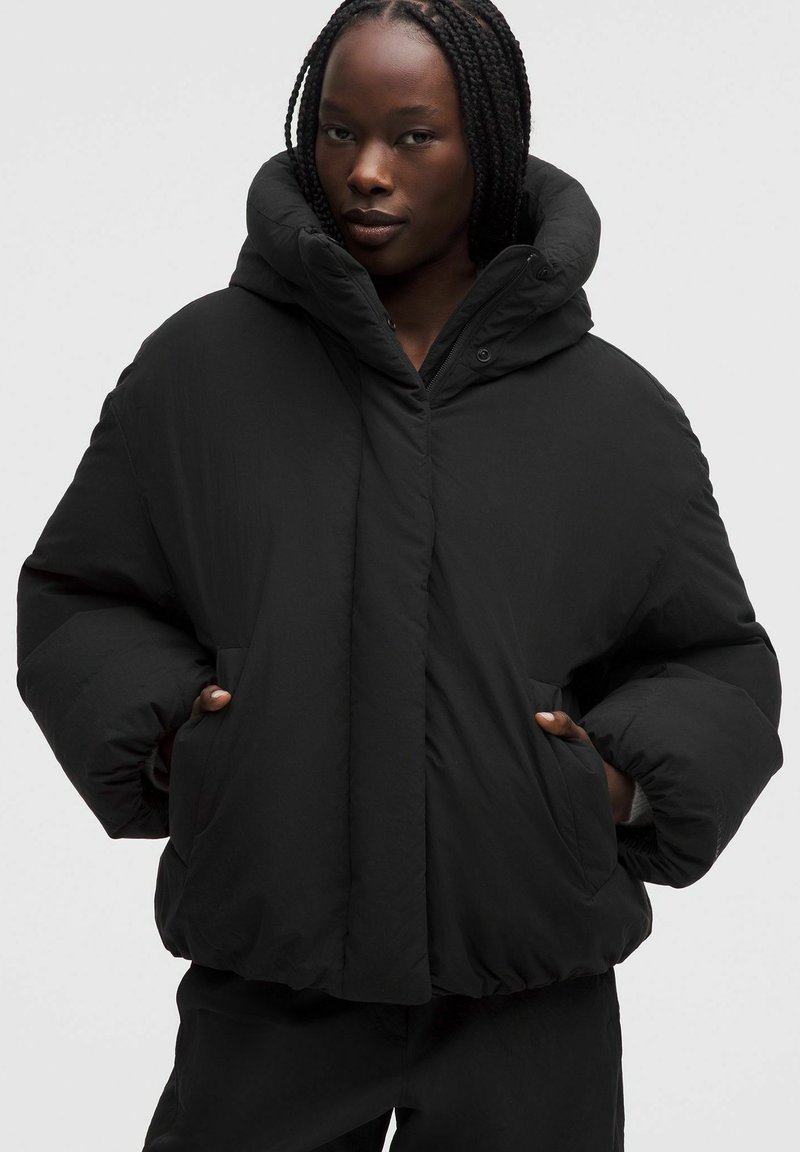 Sort pufferjakke med høj krave og stor hætte. Har en lynlåslukning, sidelommer og en afslappet pasform. Glat, mat tekstur.