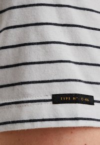 Tissu en coton à rayures blanches avec des lignes horizontales bleu marine. Il présente une étiquette noire avec un texte doré indiquant "TYPE N° : C-46" sur l'ourlet.
