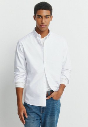 Junger Mann mit kurzen lockigen Haaren, trägt ein aufgerolltes weißes Hemd über einem weißen T-Shirt und blaue Jeans, steht vor einem schlichten hellen Hintergrund.